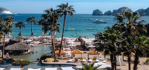 Picture of a beautiful beach resort in Los Cabos, Mexico.  The picture shows a marina, palm trees, a harbor, blue water and rocks rising from the water.