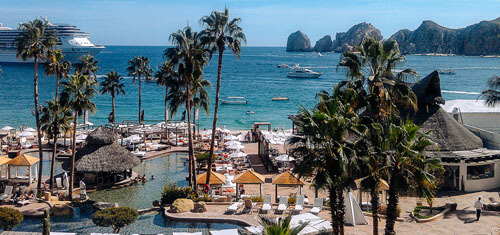 Picture of a beautiful beach and harbor in Cabo San Lucas, Mexico.  The picture shows, palm trees, boats in the harbor, blue skies and blue water.