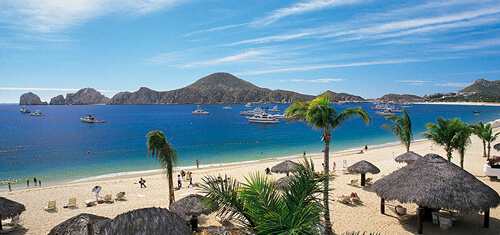 Picture of a beautiful sandy beach in Cabo San Lucas, Mexico. The picture shows small boats near the shore line and grass huts on the beach.