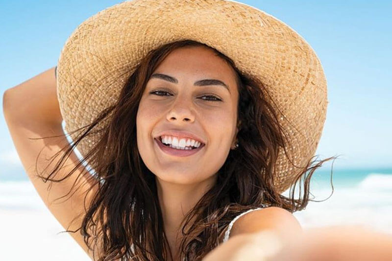 Picture of a on the beach, happy with her plastic surgery at Cabo MedVentures.  The woman is wearing tan sun hat and sitting on a Cabo beach.
