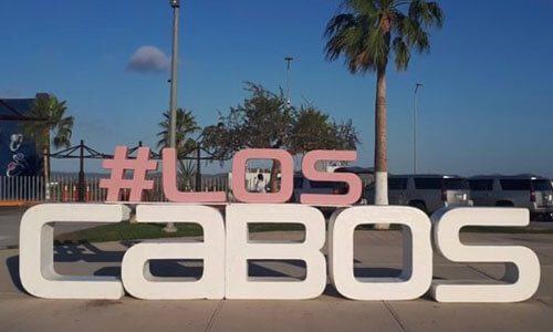 Picture of a street sign with the words “Los Cabos” in beautiful Cabo San Lucas, Mexico.  The top word “Los” is in pink color, and the bottom word “Cabos” is in white color.