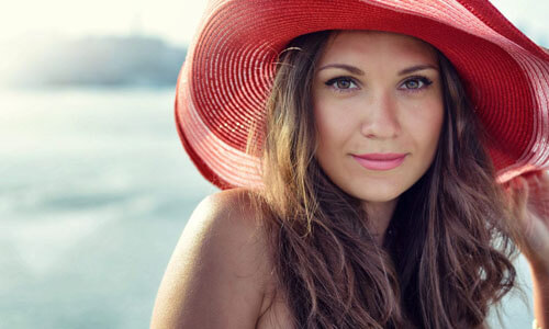 Portrait picture of a beautiful woman, happy with her face lift with neck lift procedure i in Cabo San Lucas, Mexico.  The woman has long dark hair, is looking directly into the camera and is wearing a soft red beach hat highlighting her face lift with neck lift.