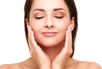 Picture of a woman with long dark brown hair, facing the camera with eyes closed, holding her hands to the side of the cheeks and happy with her perfect face lift with neck lift procedure she had with top plastic surgeons in beautiful Cabo San Lucas, Mexico.
