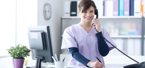 Picture of a receptionist representing Top Plastic Surgeons in beautiful Cabo San Lucas, Mexico.  The woman has short brown hair, is wearing a hospital smock and is standing at the receptionist desk while smiling at the camera